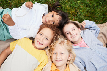 Fototapeta premium Top view portrait of diverse group of kids lying on grass in park and looking at camera smiling