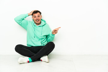 Young handsome caucasian man sitting on the floor surprised and pointing finger to the side