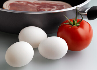 Eggs and tomato resting next to a frying pan and bacon cooking in the frying pan for breakfast