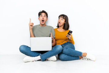 Young couple with a laptop and mobile sitting on the floor thinking an idea pointing the finger up