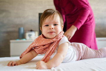 Fototapeta premium Caucasian baby lying on the bed at home in the bright room on the belly down while her mother is standing behind looking for clothes to change real people childhood and family concept