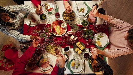 Friends holding hands and praying before dinner top view, new year tradition, sitting at dining table. International young people eating delicious food during christmas party at home. 
