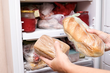A hand putting two loafs of wheat and brown bread in reserve on a shelf of a home freezer, long life food storage concept - obrazy, fototapety, plakaty
