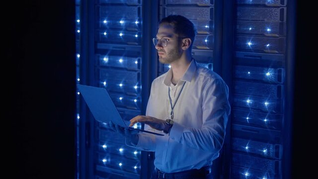 Male Server Engineer in Data Center. IT engineer inspecting a secure server cabinet using modern technology laptop coworking in data center.