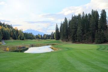 A beautiful view of of a fairway looking downhill towards a green with a lake beside it, on a beautiful golf course with autumn colours, in the rocky mountains near Canmore, Alberta, Canada - obrazy, fototapety, plakaty