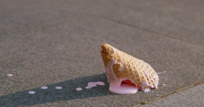 Drops of melting fruit ice cream fall on asphalt illuminated by warm rays of the summer sun, and then the ice cream itself in waffle cone falls to the ground, close up.