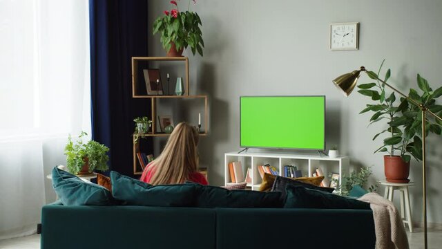 Woman watching television with green screen. Unrecognizable female person switching channels on tv with chroma key, sitting on sofa in living room. Spare time at home, relaxing and leisure.