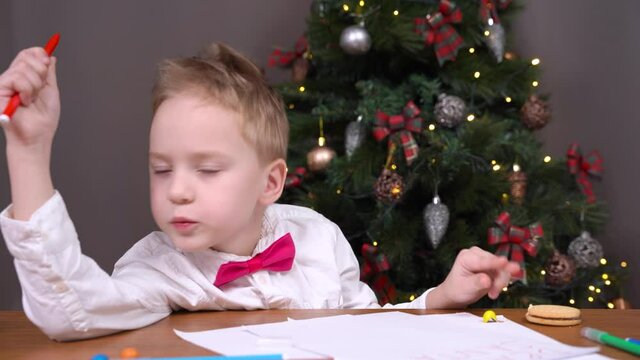 Happy little boy kid with bow tie writes letter TO Santa Claus sitting at table near decorated Christmas tree in room closeup,and waving his arms