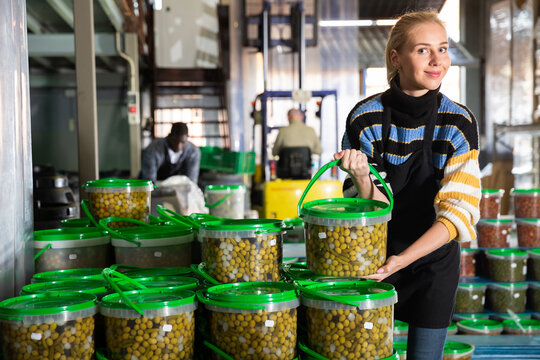 Positive girl working in food producing factory, arranging plastic buckets with pickled olives - Powered by Adobe