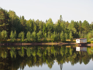 Reflection of the pine forest in the water. Old house on the shore. Morning sun. Landscape