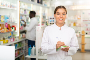 Female pharmacist writing something in notebook. Her colleague standing behind.