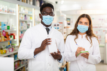 Pharmacists in face masks and white gowns standing in salesroom of drugstore