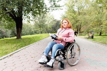 girl teenager disabled sitting in a park in a wheelchair reading a book. look at the camera. A full life for people with disabilities. Reading books and walking in the park. Outside home.

