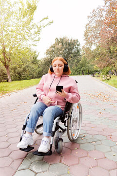 A Teenage Girl In A Wheelchair Listens To Music With Headphones And Holds A Smartphone In Her Hands While Walking In The Park. Online Communication. Love For Music. 
