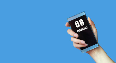 february 08. 08th day of the month, calendar date.Woman's hand holds mobile phone with blank screen on blue isolated background.Winter month, day of the year concept