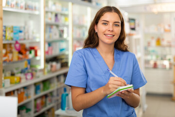 Young smiling female pharmacist working in a pharmacy is standing in the trading floor, making important notes in a notebook