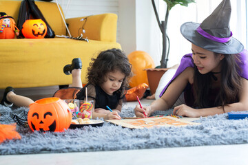 Fototapeta premium Mother and daughter in witch costumes lying on the floor and prepare decorations for Halloween party at home, little girl holding brush painting on cardboard, happy halloween together