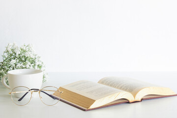 Open book with glasses, coffee and white flower over the white background with nature light.