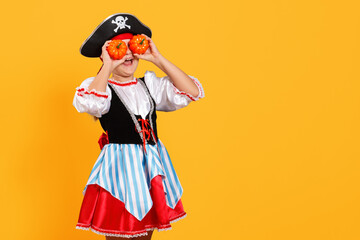Celebrating Halloween. Cheerful girl in a carnival costume of a pirate in the studio on a yellow background. The child holds the pumpkins near his eyes.