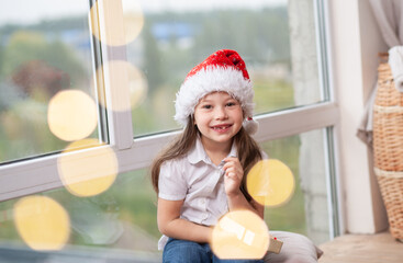 A cute little girl in a Santa Claus hat holds a gift in her hands, around special defocused glare...