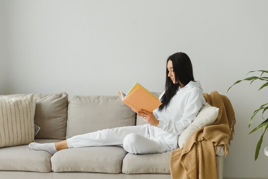 Pretty young woman enjoying reading a book at home lying on the sofa smiling in pleasure in casual clothing