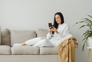 Portrait of a cheerful young woman using mobile phone while relaxing on a couch at home - Powered by Adobe
