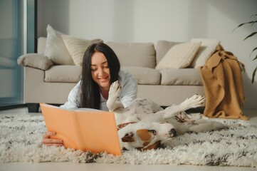 Woman in cozy home clothes relaxing at home with dog reading a book. Comfy lifestyle.