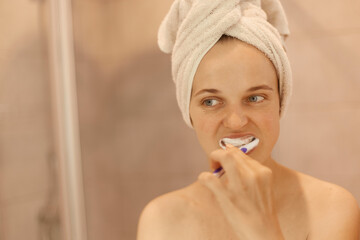 Portrait of attractive young adult woman with perfect skin with towel on her head brushing her teeth, hygiene procedures at home in bathroom, holding toothbrush in hands.