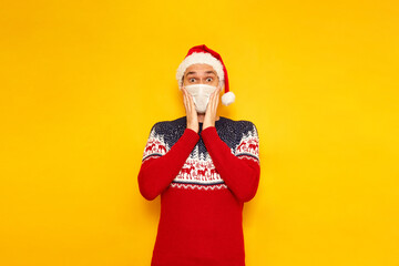 shocked, excited man in Christmas sweater with reindeer, medical mask, New Year's hat, Santa Claus hat, holds his hands to his face, wow effect. isolated yellow background with space for text. holiday
