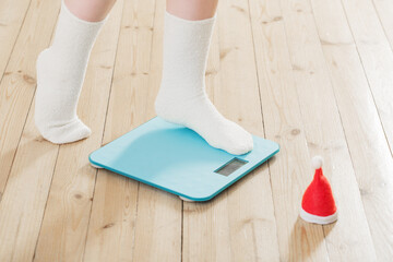 Female feet standing on blue electronic scales for weight control  with Christmas santa hat on wooden floor