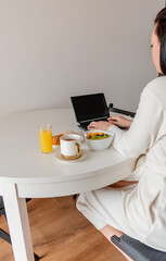 Fototapeta na wymiar Young woman in comfortable white robe works on laptop while sitting at table the kitchen. Work at home for breakfast.