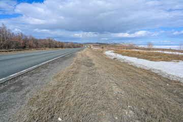 Highway road in the steppes, snow, bushes, grass and cloudy sky. Volga River. Spring landscape.