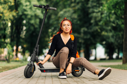 Young asian woman, wearing glasses riding electric scooter in the city, full length portrait of a woman