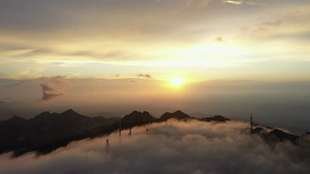 View from above, flying above the clouds, stunning aerial view of a mountain range with an antenna farm surrounded by clouds during a beautiful sunrise. Mount Limbara (Monte Limbara) Sardinia, Italy.
