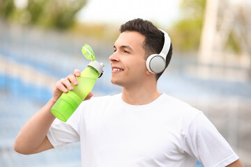 Sporty young man in headphones drinking water at stadium