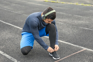 Muscular young man in headphones tying shoe lace at stadium