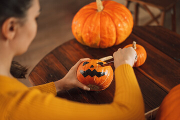 Girl in an orange sweater cuts an evil face on pumpkin with knife.