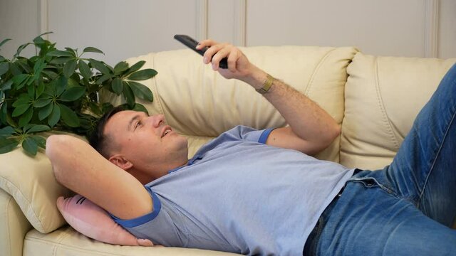 happy young man lies on sofa and texting on phone. Indoor shot of smiling millennial man surfing internet on mobile phone, messaging friends online and checking newsfeed via social media