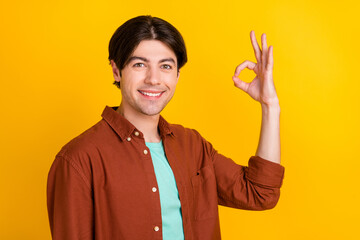 Photo of excited funny young man wear brown shirt smiling showing okey isolated yellow color background