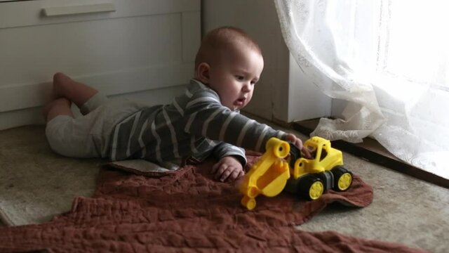 Cute caucasian baby learning to crawl and play on the floor with a plastic yellow tractor, toddler 5 6 months on the stomach on the floor, children development, subject manipulative activity