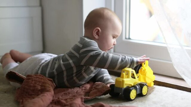 Cute caucasian baby learning to crawl and play on the floor with a plastic yellow tractor, toddler 5 6 months on the stomach on the floor, children development, subject manipulative activity