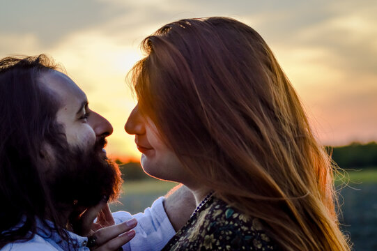 Backlight portrait of a young couple standing to kiss with sunset, he has a beard and long hair, her long red hair