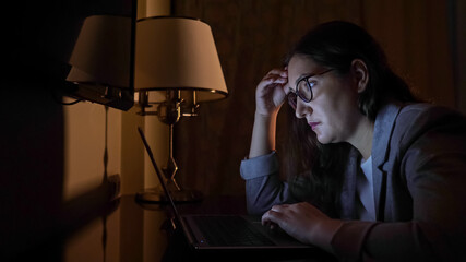 woman in a business suit sits at a laptop on a dark evening.