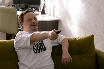 Guy with down syndrome watching TV at home, using TV remote.Man sitting on th green sofa at the room.Empty space