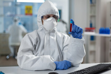 Chemist researcher with protective suit against covid19 holding medical vacutainer analyzing blood results during coronavirus vaccine development. Scientist woman working in hospital lab