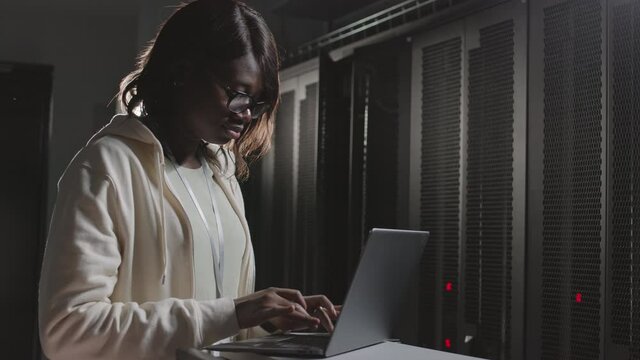 Waist-up of young African American female programmer wearing eyeglasses, using portable computer, typing in dark server room