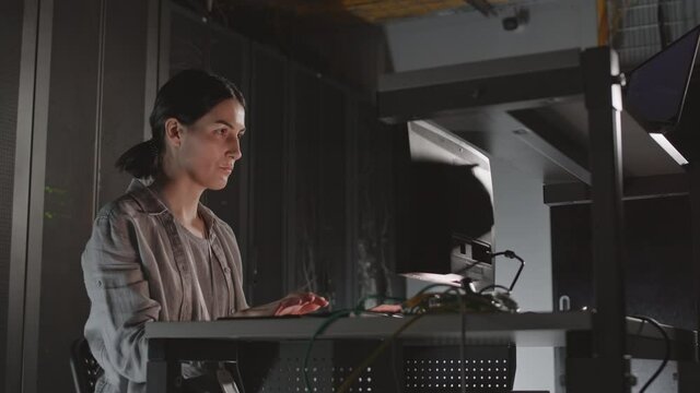 Low angle medium of black-haired Caucasian woman sitting at desk in server room, using computer, typing on keyboard