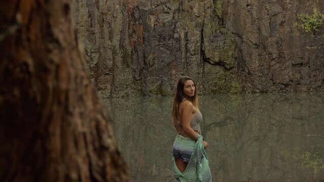 Young Caucasian Woman Standing Against Natural Waterhole Turn And Smile As The Camera Pass By. - slide shot