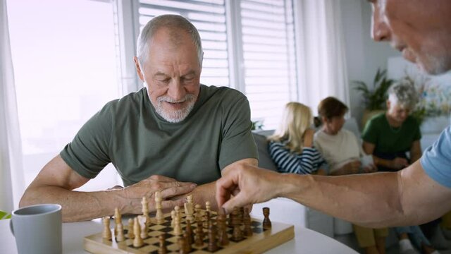 Two senior friends playing chess indoors, social gathering concept.