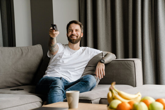 Bearded european man smiling and watching TV while sitting on sofa